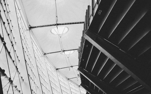 A black and white architectural photograph featuring geometric structures, including a modern roof with fabric stretched across metal beams and an angular ceiling design. The image captures the contrast between light streaming through translucent surfaces and dark shadow areas.