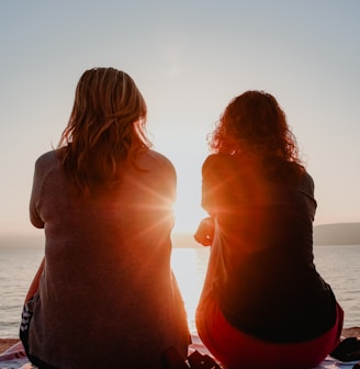 two woman sitting on beach sand while facing sunlight