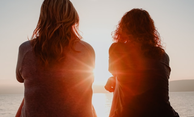 two woman sitting on beach sand while facing sunlight