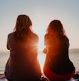 two woman sitting on beach sand while facing sunlight