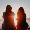 two woman sitting on beach sand while facing sunlight