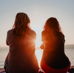 two woman sitting on beach sand while facing sunlight