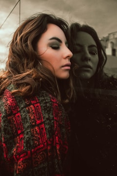A woman with closed eyes leans against a reflective surface, creating a calm and introspective mood. She is wearing a patterned red and dark sweater, and the background shows an overcast sky, enhancing the reflective atmosphere.