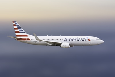A large commercial airplane with the American logo is flying through an open sky with soft gradient hues ranging from light blue to gray.
