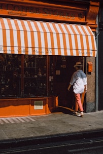 A person wearing a hat and white shirt is entering a store with orange and white striped awning. The store is called Algerian Coffee Stores Ltd, established in 1887. The storefront features an orange facade with a glass display window.