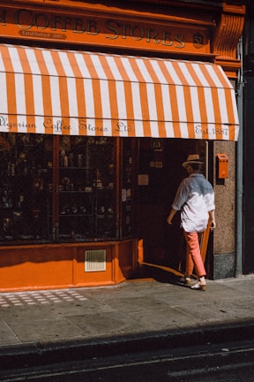 A person wearing a hat and white shirt is entering a store with orange and white striped awning. The store is called Algerian Coffee Stores Ltd, established in 1887. The storefront features an orange facade with a glass display window.
