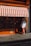 A person wearing a hat and white shirt is entering a store with orange and white striped awning. The store is called Algerian Coffee Stores Ltd, established in 1887. The storefront features an orange facade with a glass display window.