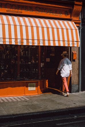 A person wearing a hat and white shirt is entering a store with orange and white striped awning. The store is called Algerian Coffee Stores Ltd, established in 1887. The storefront features an orange facade with a glass display window.