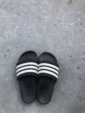 Black slide sandals with three white stripes across the top are placed on a rough gray concrete surface. The brand name is subtly embossed on the footbed of each sandal.
