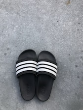 Black slide sandals with three white stripes across the top are placed on a rough gray concrete surface. The brand name is subtly embossed on the footbed of each sandal.