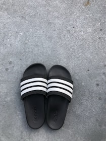 Black slide sandals with three white stripes across the top are placed on a rough gray concrete surface. The brand name is subtly embossed on the footbed of each sandal.