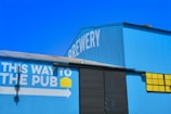 A large blue building with the word 'Brewery' written on it in white letters. There's a sign on the side of the building with the text 'This way to the pub' in white, accompanied by a yellow square icon and a white arrow pointing right. The building features corrugated metal siding and large black doors, with a section of bright yellow windows.