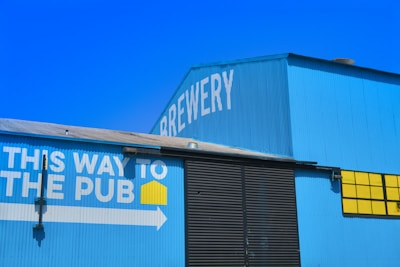 A large blue building with the word 'Brewery' written on it in white letters. There's a sign on the side of the building with the text 'This way to the pub' in white, accompanied by a yellow square icon and a white arrow pointing right. The building features corrugated metal siding and large black doors, with a section of bright yellow windows.