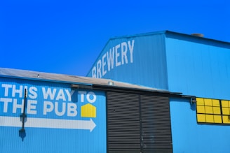 A large blue building with the word 'Brewery' written on it in white letters. There's a sign on the side of the building with the text 'This way to the pub' in white, accompanied by a yellow square icon and a white arrow pointing right. The building features corrugated metal siding and large black doors, with a section of bright yellow windows.