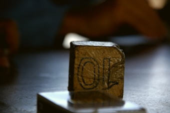 A small, rectangular package wrapped in a brown material sits on a surface. The package has markings or writing on it, and another object, possibly a shiny box or scale, is partially visible in the foreground.