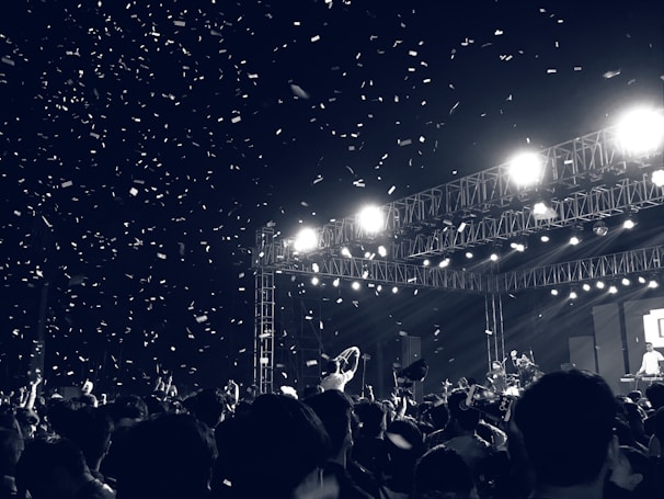 A large crowd is gathered at an outdoor concert during nighttime, with many people raising their hands and recording the event. Bright stage lights illuminate the scene, and there is confetti falling through the air.