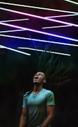 A person with long dreadlocks appears to be in motion with their head tilted back and mouth open, possibly singing or shouting. Above them are colorful diagonal neon lights creating an energetic and vibrant atmosphere.