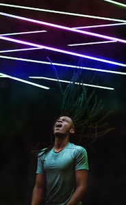 A person with long dreadlocks appears to be in motion with their head tilted back and mouth open, possibly singing or shouting. Above them are colorful diagonal neon lights creating an energetic and vibrant atmosphere.