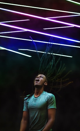 A person with long dreadlocks appears to be in motion with their head tilted back and mouth open, possibly singing or shouting. Above them are colorful diagonal neon lights creating an energetic and vibrant atmosphere.
