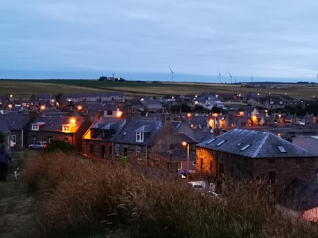 A quaint village scene during twilight, featuring stone houses with warm glowing windows and street lamps. The village is nestled in a vast, lush landscape with wind turbines dotting the horizon. Grassy fields are in the foreground with a person and a dog walking.