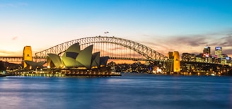 The iconic Sydney Opera House stands prominently against the backdrop of the Sydney Harbour Bridge at sunset. The city skyline is illuminated with twinkling lights as the sky transitions through shades of blue, orange, and pink. Calm waters of the harbor reflect the lights and colors of the scene.