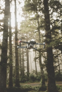 A drone is hovering in front of a dense forest with tall trees and ambient sunlight filtering through the branches. The background is slightly foggy, creating a serene and mysterious atmosphere.