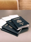 Five dark blue passports from the United States of America are placed on a gray table. Each passport displays the country's emblem and name in gold lettering. In the background, there is a chair with a circular patterned fabric.