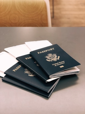 Five dark blue passports from the United States of America are placed on a gray table. Each passport displays the country's emblem and name in gold lettering. In the background, there is a chair with a circular patterned fabric.