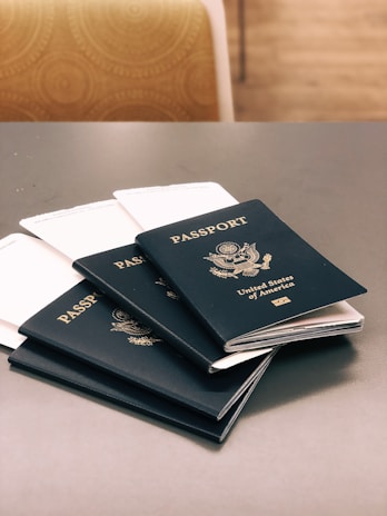Five dark blue passports from the United States of America are placed on a gray table. Each passport displays the country's emblem and name in gold lettering. In the background, there is a chair with a circular patterned fabric.