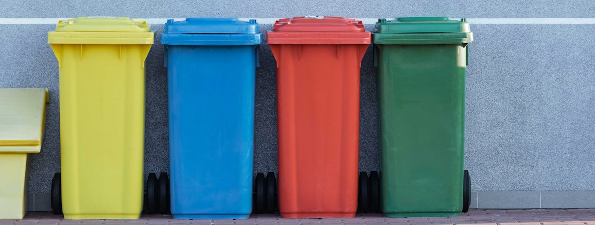 four assorted-color trash bins beside gray wall