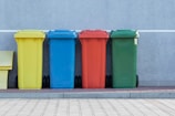 four assorted-color trash bins beside gray wall
