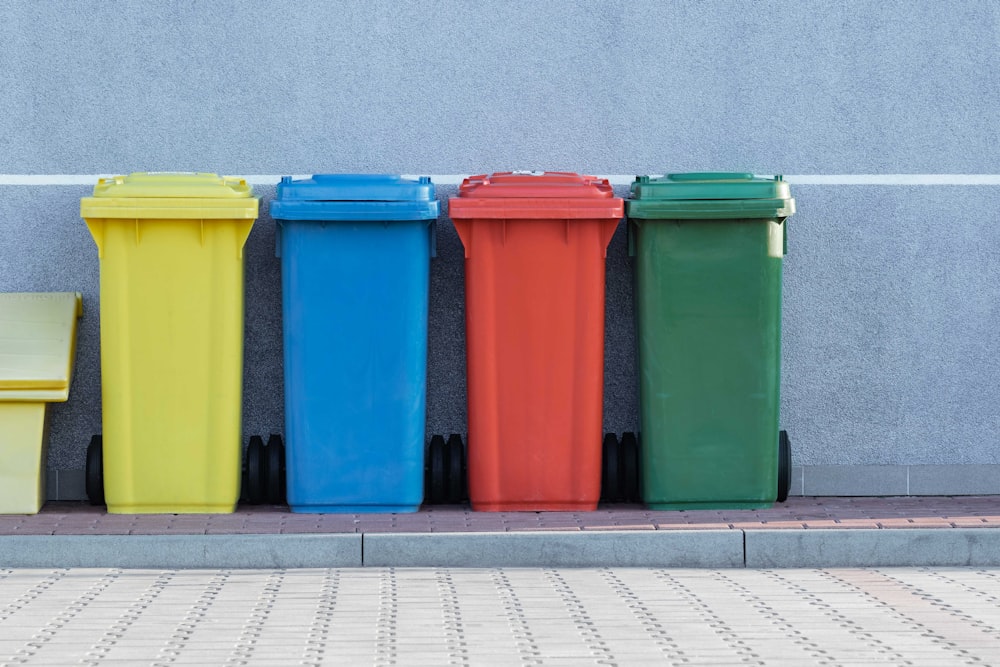 Cuatro cubos de basura de colores variados junto a la pared gris