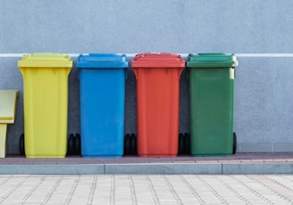 four assorted-color trash bins beside gray wall