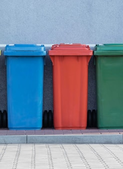 four assorted-color trash bins beside gray wall