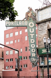 A vintage sign for the Portland Outdoor Store featuring a cowboy riding a horse. The sign is attached to a corner building with an old-style brick facade. A modern, reddish-brown building with multiple windows is seen in the background. A streetlamp and traffic signal are visible on the left side.