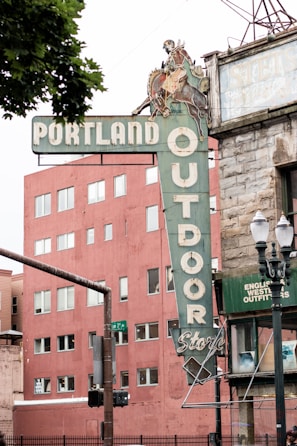 A vintage sign for the Portland Outdoor Store featuring a cowboy riding a horse. The sign is attached to a corner building with an old-style brick facade. A modern, reddish-brown building with multiple windows is seen in the background. A streetlamp and traffic signal are visible on the left side.