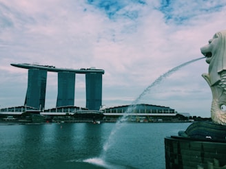 A waterfront scene featuring a large iconic structure with three towers topped by a boat-shaped platform, known as Marina Bay Sands. In the foreground is a large Merlion statue spouting water into the bay. The sky is overcast with scattered clouds.