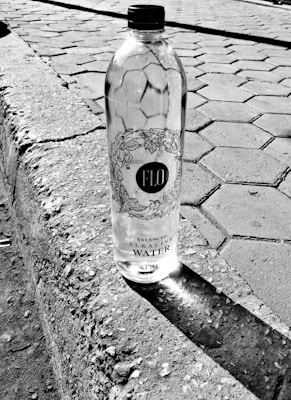 Plastic bottle of FLO balanced alkaline water placed on a curb made of concrete with hexagonal tiled pavement in the background. The bottle casts a shadow to its side.