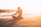 A person is seated in a meditative pose on a sandy beach during sunset, with the ocean waves gently rolling in the background. The golden light of the setting sun creates a serene and peaceful atmosphere, casting a long shadow on the sand. Another person is visible swimming in the water further away.