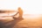 A person is seated in a meditative pose on a sandy beach during sunset, with the ocean waves gently rolling in the background. The golden light of the setting sun creates a serene and peaceful atmosphere, casting a long shadow on the sand. Another person is visible swimming in the water further away.