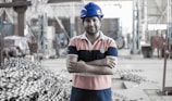 A person wearing a blue hard hat and a striped polo shirt stands confidently with arms crossed in an industrial setting. The background shows a large workshop area with metal equipment and structures.