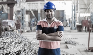 A person wearing a blue hard hat and a striped polo shirt stands confidently with arms crossed in an industrial setting. The background shows a large workshop area with metal equipment and structures.