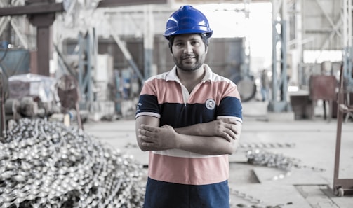 A person wearing a blue hard hat and a striped polo shirt stands confidently with arms crossed in an industrial setting. The background shows a large workshop area with metal equipment and structures.