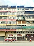 A multi-story building with a weathered facade is visible. The structure features a variety of window styles and colors, indicating a blend of residential and commercial uses. The paint on the building appears faded and peeling, adding a sense of age to the architecture. On the ground floor, signage with Chinese characters and a sign for 'Richfield Realty' identify businesses. A red and white taxi is parked in front of the building, and traffic cones are placed along the sidewalk. The overall atmosphere is urban and slightly rundown.