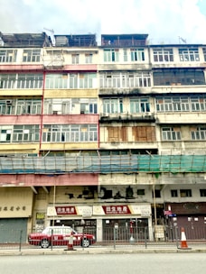 A multi-story building with a weathered facade is visible. The structure features a variety of window styles and colors, indicating a blend of residential and commercial uses. The paint on the building appears faded and peeling, adding a sense of age to the architecture. On the ground floor, signage with Chinese characters and a sign for 'Richfield Realty' identify businesses. A red and white taxi is parked in front of the building, and traffic cones are placed along the sidewalk. The overall atmosphere is urban and slightly rundown.