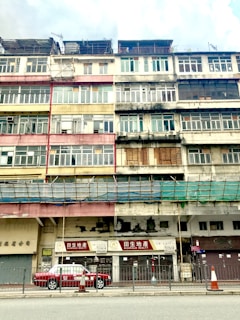 A multi-story building with a weathered facade is visible. The structure features a variety of window styles and colors, indicating a blend of residential and commercial uses. The paint on the building appears faded and peeling, adding a sense of age to the architecture. On the ground floor, signage with Chinese characters and a sign for 'Richfield Realty' identify businesses. A red and white taxi is parked in front of the building, and traffic cones are placed along the sidewalk. The overall atmosphere is urban and slightly rundown.