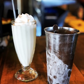A creamy milkshake topped with whipped cream sits in a tall glass with a straw, next to a stainless steel cup on a wooden table.