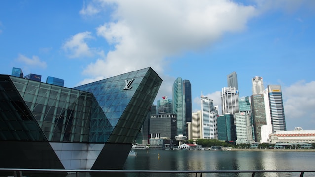 The image displays a cityscape with modern, high-rise buildings and a distinctive glass structure on the waterfront. The skyline features various commercial skyscrapers with diverse architectural designs, set against a blue sky with scattered clouds.
