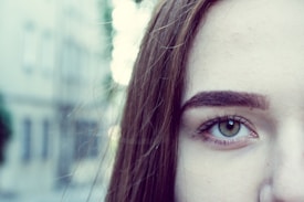 A close-up of a person's face focusing primarily on one eye. The image captures a clear view of a green eye with well-defined eyebrows and long eyelashes. The background is blurred, highlighting the subject's face.