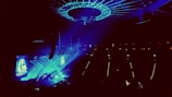 A concert venue illuminated with vibrant blue and purple lights, showcasing a large stage with performers. An overhead circular structure is present, adding to the modern ambiance. The audience is dimly visible, focusing their attention on the performance.